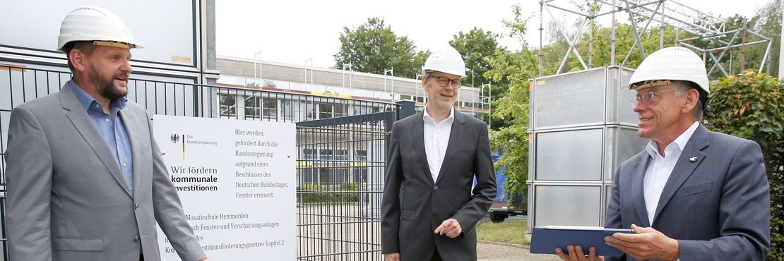 Schulleiter Markus Steinhauer, Projektleiter Franz Steves und Landrat Hans-Jürgen Petrauschke  mit Schutzhelmen stehen mit Abstand vor einem Baustellenschild