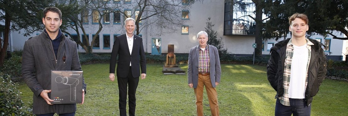 Die in der Bildbeschreibung genannten Personen stehen mit Abstand zueinander vor einem Gebäude. Trainer Francisco Frederico hält einen Karton mit einem Bild der Massagepistole in der Hand. 