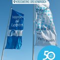 Das Bild zeigt zwei Fahnenmasten mit je einer Fahne vor einem blauen Himmel.  Die linke Fahne ist blau und trägt den Slogan „Wasser ist Leben“ sowie ein „Trinkwasser“-Logo. Die rechte Fahne ist weiß mit blauen Punkten und der Aufschrift „Qualität für Vertrauen“.  Im unteren Bereich ist das Logo der Kreiswerke Grevenbroich mit dem Jubiläumslogo „50 Jahre | 50 Orte“ zu sehen.  Das Bild suggeriert eine Werbekampagne oder ein Jubiläum dieser Wasserwerke im Rhein-Kreis Neuss. © Kreiswerke Grevenbroich GmbH