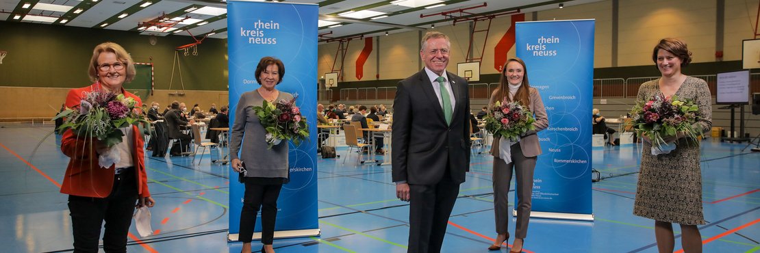 Die in der Beschreibung genannten Personen stehen mit Abständen in einer großen Turnhalle, die Damen haben je einen Strauss Blumen in der Hand. Im Hintergrund sieht man Kreistagsabgeordnete an Ihren Plätzen sitzen. 