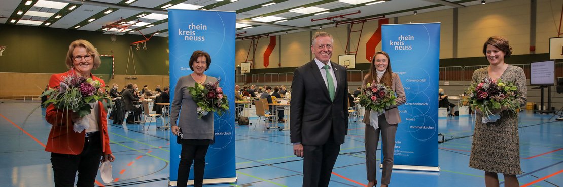 Die in der Beschreibung genannten Personen stehen mit Abständen in einer großen Turnhalle, die Damen haben je einen Strauss Blumen in der Hand. Im Hintergrund sieht man Kreistagsabgeordnete an Ihren Plätzen sitzen. 