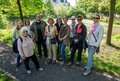 Das Foto zeigt eine Gruppe von etwa zehn Erwachsenen, die sich in einem Park neben einem Fluss versammelt haben. Sie scheinen eine Führung oder einen Ausflug zu machen, da einige von ihnen einen Flyer oder eine Broschüre in der Hand halten. Im Hintergrund ist ein Gebäude im Stil eines Herrenhauses zu sehen, und die Atmosphäre wirkt entspannt und freundlich.