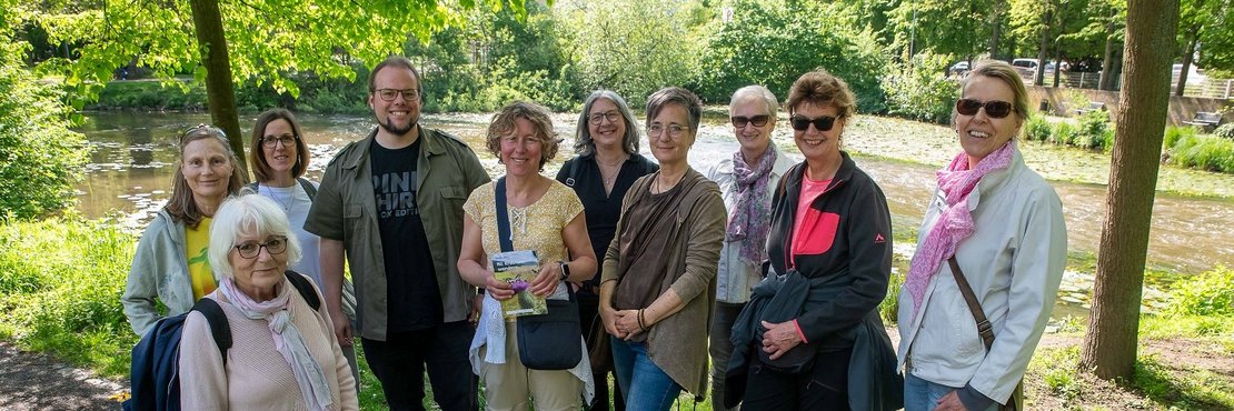 Das Foto zeigt eine Gruppe von etwa zehn Erwachsenen, die sich in einem Park neben einem Fluss versammelt haben. Sie scheinen eine Führung oder einen Ausflug zu machen, da einige von ihnen einen Flyer oder eine Broschüre in der Hand halten. Im Hintergrund ist ein Gebäude im Stil eines Herrenhauses zu sehen, und die Atmosphäre wirkt entspannt und freundlich.