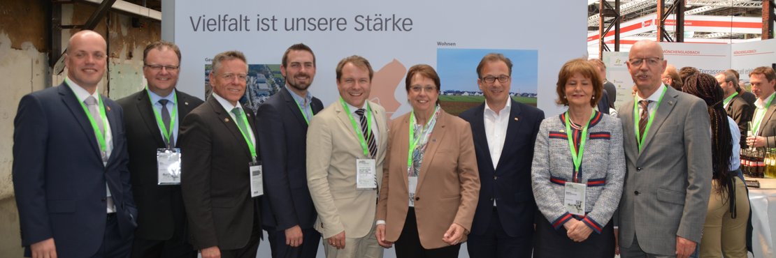 Landrat Hans-Jürgen Petrauschke mit den Bürgermeisterinnen und Bürgermeistern aus dem Rhein-Kreis Neuss vor einer Werbestellwand in einer Halle