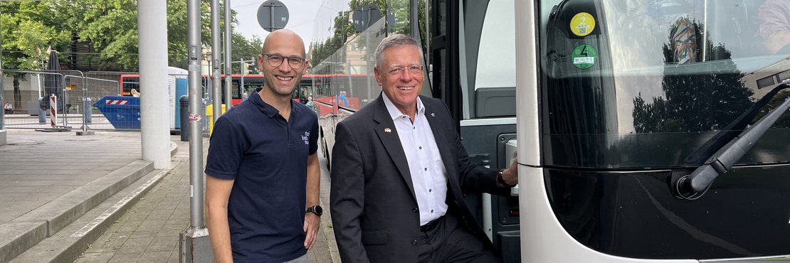Zwei männliche Personen stehen an der Eingangstüre eines weißen Reisebusses und lächeln.