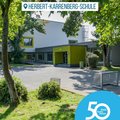 Das Bild zeigt die Herbert-Karrenberg-Schule. Es ist ein Foto des Schulgebäudes von außen, das einen modernen Stil mit hellen Farben (weiß und grün) aufweist.  Im Vordergrund befindet sich ein Teil des Schulhofs mit Rasenflächen und Bäumen. Rechts unten ist ein Logo mit der Aufschrift "50 Jahre | 50 Orte" zu sehen, das auf ein Jubiläum hinweist.  Ein Fahrrad steht links im Bild. © Rhein-Kreis Neuss