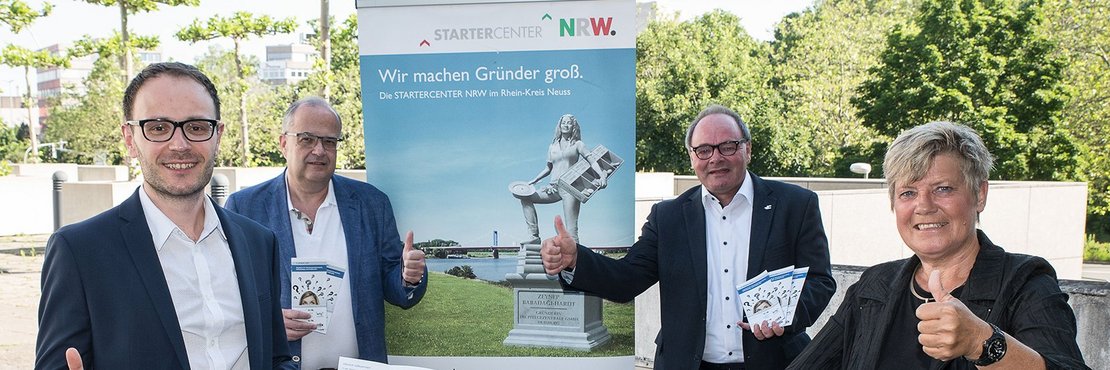Die Personen stehen vor einen Banner mit der Aufschrift: Startercenter NRW - Wir machen Gründer groß. Sie halten Faltblätter in den Händen. 