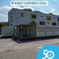 Das Bild zeigt die Herbert-Karrenberg-Schule an einem sonnigen Tag. Im Vordergrund stehen mehrere Container, die vermutlich als zusätzliche Klassenräume oder Aufenthaltsbereiche dienen. Das Hauptgebäude der Schule ist ein moderner, zweistöckiger Bau mit farbigen Akzenten an den Fenstern.  Ein Logo mit zwei stilisierten, sich haltenden Figuren ist an der Fassade angebracht. Im unteren rechten Bildbereich befindet sich ein Jubiläumslogo, das auf 50 Jahre und 50 Orte im Rhein-Kreis Neuss hinweist.  Das Gesamtbild vermittelt einen Eindruck von einer modernen Schule. © Rhein-Kreis Neuss