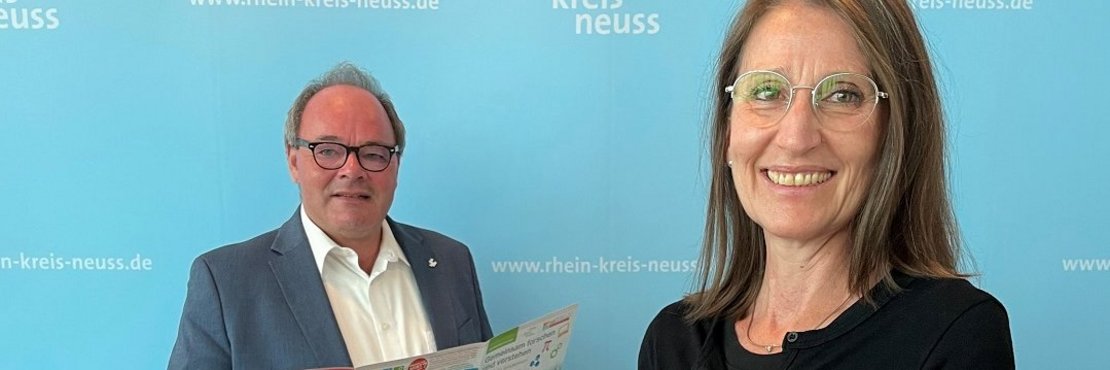 Zwei Personen mit Flyern vor einer Pressewand.