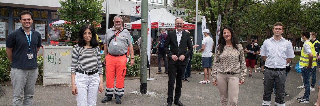 Genannte Personen stehen versetzt mit Abständen auf dem Bürgersteig vor dem Bürgerhaus Erfttal. Im Hintergrund vor dem Eingang eine schlange wartender Menschen und Hilfskräfte vor dem Eingang. 