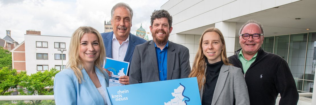 Zwei Frauen und drei Männer halten ein blaues Plakat in der Hand und lächeln in die Kamera.