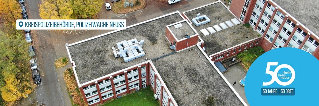 Das Bild zeigt eine Luftaufnahme der Kreispolizeibehörde, Polizeiwache Neuss.  Im Vordergrund ist ein Teil des Gebäudes mit dem Dach zu sehen, auf dem sich diverse technische Anlagen befinden.  Im Hintergrund sind Parkplätze mit Autos und die umliegende herbstliche Landschaft mit gelb-braunen Bäumen sichtbar. Rechts unten im Bild befindet sich ein blaues Logo, das auf das 50-jährige Jubiläum des Rhein-Kreises Neuss hinweist.