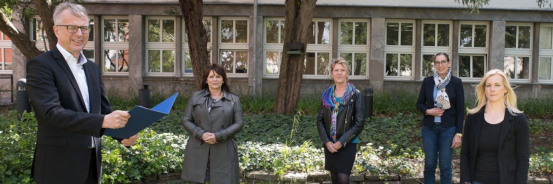 Genannte Personen stehen auf Blumenwiese. Im Hintergrund Bäume und ein Gebäude.