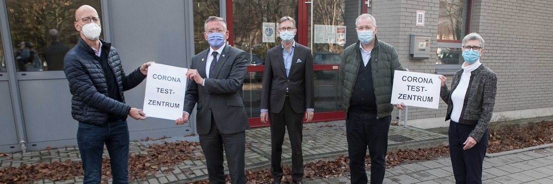 Die in der Bildbeschreibung genannten Personen stehen mit Mund-Nase-Masken vor dem Gebäude des Testezentrums. Sie halten Schilder hoch mit der Aufschrift: Corona Test-Zentrum