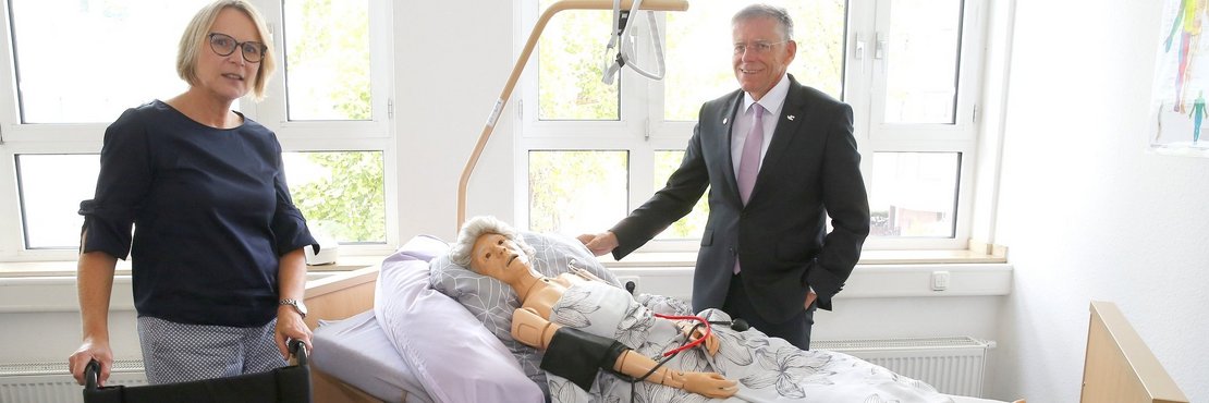 Landrat Hans-Jürgen Petrauschke steht an einem Krankenbett indem eine Pflegepuppe leigt. Frau Groth steht vor dem Bett, hinter einem leeren Rollstuhl