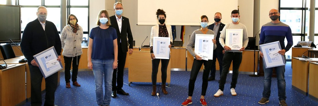 Die in der Bildbeschreibung genannten Personen stehen in einem Raum. Sie tragen Mund-Nasenschutz und halten Ihre Urkunden.