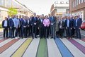 Das Bild zeigt eine große Gruppe von Menschen, die auf einem regenbogenfarbenen Zebrastreifen stehen und für ein Gruppenfoto posieren. Die Gruppe besteht überwiegend aus Männern und Frauen in Businesskleidung. Im Vordergrund stehen einige Personen, die etwas prominenter zu sehen sind als der Rest der Gruppe. Der Hintergrund besteht aus Industriegebäuden.  Das Bild wirkt formell und könnte einen offiziellen Anlass darstellen.