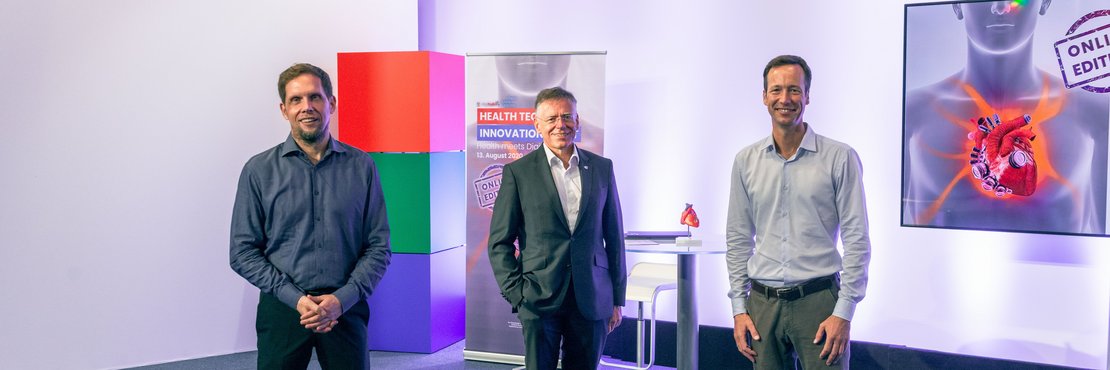 Landrat Hans-Jürgen Petrauschke steht mit zwei Herren vor einem modernen Edelstahl-Stehtisch mit Glasplatte. Im Hintergrund sieht man drei große aufeinander gestapelte  Quader in rot, grün und blau