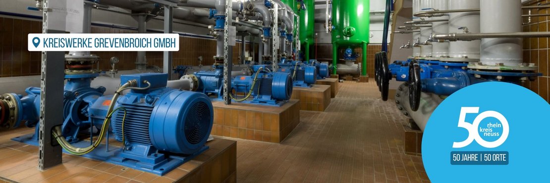 Das Bild zeigt eine Maschinenhalle mit mehreren blauen Pumpen und einem komplexen System aus grauen Rohren und Ventilen. Die Pumpen stehen auf quadratischen Sockeln aus braunen Fliesen. Im Hintergrund befinden sich zwei große grüne Behälter. Die gesamte Szenerie wirkt industriell und technisch.  Das Bild fokussiert auf die technische Anlage und wirkt sauber und ordentlich.