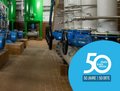 Das Bild zeigt eine Maschinenhalle mit mehreren blauen Pumpen und einem komplexen System aus grauen Rohren und Ventilen. Die Pumpen stehen auf quadratischen Sockeln aus braunen Fliesen. Im Hintergrund befinden sich zwei große grüne Behälter. Die gesamte Szenerie wirkt industriell und technisch.  Das Bild fokussiert auf die technische Anlage und wirkt sauber und ordentlich.
