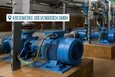 Das Bild zeigt eine Maschinenhalle mit mehreren blauen Pumpen und einem komplexen System aus grauen Rohren und Ventilen. Die Pumpen stehen auf quadratischen Sockeln aus braunen Fliesen. Im Hintergrund befinden sich zwei große grüne Behälter. Die gesamte Szenerie wirkt industriell und technisch.  Das Bild fokussiert auf die technische Anlage und wirkt sauber und ordentlich.
