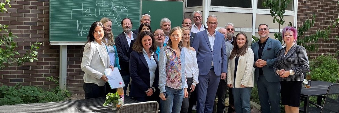 Das Bild zeigt eine Gruppe von etwa 15 Personen, die vor einer Backsteinmauer stehen.  Im Hintergrund steht eine grüne Tafel, auf der mit Kreide „Hallo“ und „Ne…“ (vermutlich unvollständig) geschrieben steht.  Die Personen wirken wie eine Gruppe von Erwachsenen, möglicherweise ein Arbeitsteam oder eine politische Gruppe, die sich für ein Foto versammelt haben.  Manche halten Dokumente oder Getränke.  Der Ort scheint ein Außenbereich zu sein, mit Gartenelementen wie Pflanzen und Tischen mit Stühlen im Vordergrund. Die Atmosphäre ist freundlich und ungezwungen.