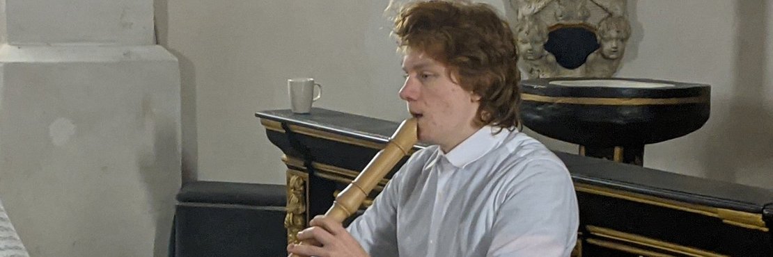 Junger Mann sitzt auf Stuhl und spielt eine große Blockflöte