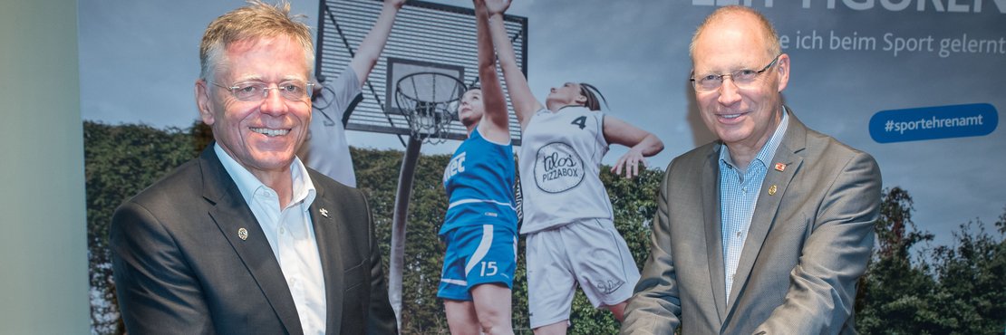 Landrat Hans-Jürgen Petrauschke überreicht ein Dokument an Dr. Hermann-Josef Baaken. Im Hintergrund eine Fotowand auf der junge Menschen Basketball spielen. 