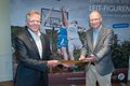 Landrat Hans-Jürgen Petrauschke überreicht ein Dokument an Dr. Hermann-Josef Baaken. Im Hintergrund eine Fotowand auf der junge Menschen Basketball spielen. 