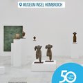 Das Bild zeigt einen Ausstellungsraum im Museum Insel Hombroich.  Im Raum stehen mehrere weiße Podesten, auf denen sich verschiedene Skulpturen befinden, die aus Stein oder ähnlichem Material gefertigt zu sein scheinen und einen asiatisch anmutenden Stil haben.  Im Hintergrund ist ein großes, grün-blaues Gemälde zu sehen.  Das Bild wird durch ein Logo mit der Aufschrift "50 Jahre | 50 Orte" und Informationen zum Museum und den Fotografen ergänzt.  Es vermittelt einen Eindruck von Ruhe und künstlerischer Atmosphäre. © Museum Insel Hombroich / 
