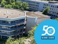 Das Bild zeigt eine Luftaufnahme des Berufskollegs für Technik und Informatik (BTI) im Rhein-Kreis Neuss.  Das Gebäude ist ein mehrstöckiger, sechseckiger Bau, umgeben von Bäumen und anderen Gebäuden. Im Bild ist auch ein Logo zu sehen, das auf das 50-jährige Jubiläum des Rhein-Kreises Neuss hinweist.