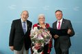 Zwei männliche Personen und weibliche Person in der Mitte mit einem Blumenstrauß