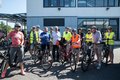 Das Bild zeigt eine Gruppe von etwa fünfzehn Personen, die vor einem modernen, hellgrauen Gebäude mit großen Fenstern stehen. Alle tragen Fahrradhelme und viele tragen auch Warnwesten. 