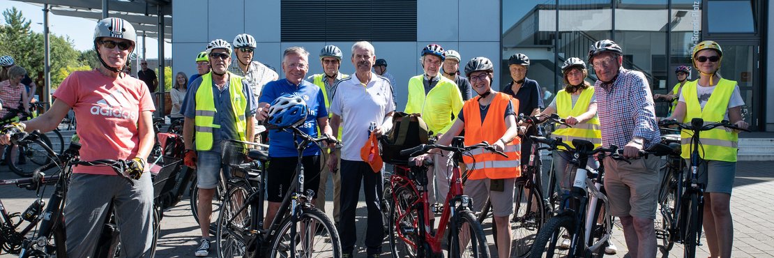 Das Bild zeigt eine Gruppe von etwa fünfzehn Personen, die vor einem modernen, hellgrauen Gebäude mit großen Fenstern stehen. Alle tragen Fahrradhelme und viele tragen auch Warnwesten. 