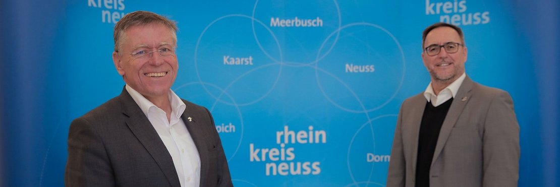 Landrat Petrauschke und Baudezernent Vieten stehen vor einer blauen Fotowand mit dem Logo des Rhein-Kreis Neuss