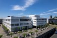 Das Bild zeigt eine Luftaufnahme des Kreishauses Neuss, einem modernen, weißen Gebäude mit einer großzügigen Vorplatzgestaltung. Der quadratische Vorplatz ist mit Bäumen in großen Pflanzgefäßen angelegt. Im Hintergrund ist ein Teil der Neusser Innenstadt mit der katholischen Kirche St. Quirinus zu sehen.  Der Himmel ist strahlend blau mit wenigen Wolken.