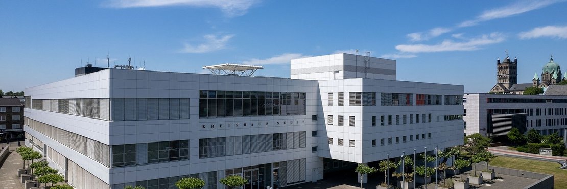 Das Bild zeigt eine Luftaufnahme des Kreishauses Neuss, einem modernen, weißen Gebäude mit einer großzügigen Vorplatzgestaltung. Der quadratische Vorplatz ist mit Bäumen in großen Pflanzgefäßen angelegt. Im Hintergrund ist ein Teil der Neusser Innenstadt mit der katholischen Kirche St. Quirinus zu sehen.  Der Himmel ist strahlend blau mit wenigen Wolken.