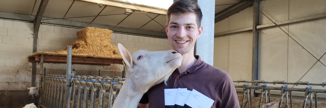 Mann neben Ziege mit Broschüre in der Hand.