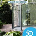 Das Bild zeigt einen Teil des Graubner Pavillons im Museum Insel Hombroich.  Es ist ein moderner, verglaster Rundbau, der in eine grüne, parkähnliche Umgebung eingebettet ist. Im Vordergrund sind Pflanzen zu sehen. Ein runder Aufkleber mit der Zahl 50 und dem Hinweis auf ein Jubiläum ("50 Jahre | 50 Orte")  des Rhein-Kreises Neuss ist ebenfalls im Bild abgebildet. © Museum Insel Hombroich / Jennifer Eckert, Erwin Heerich