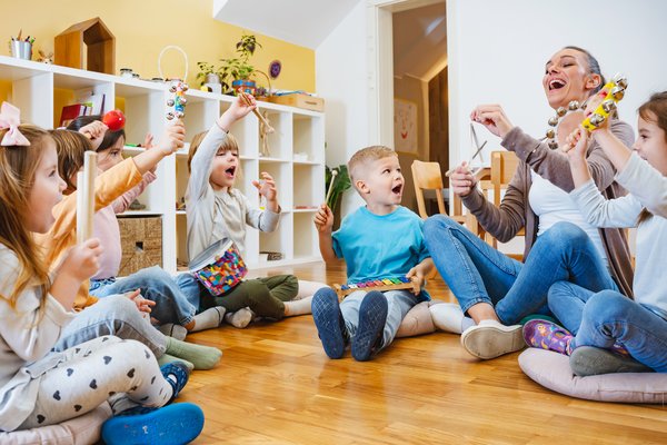 dekorativ, Spielgruppe von Kindern mit Betreuungsperson fröhlich auf dem Boden sitzend und musizierend