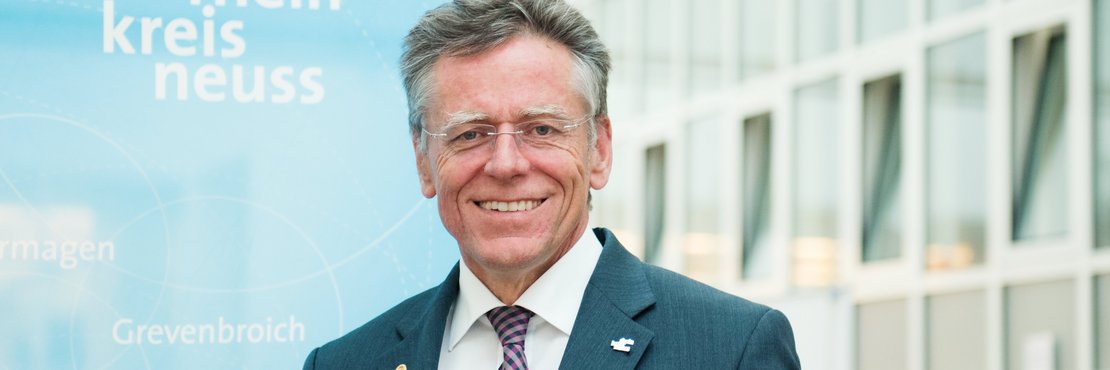 Landrat Hans-Jürgen Petrauschke steht lächelnd vor einer blauen Fahne mit weißem Kreislogo