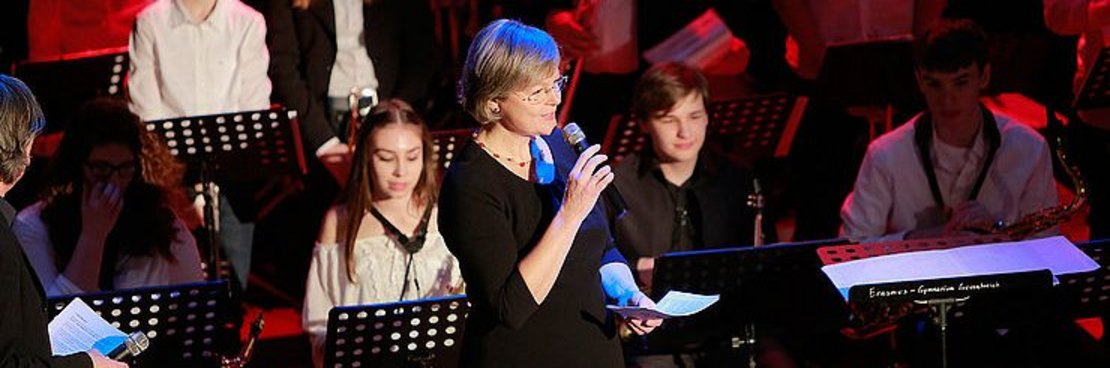 Eine Frau begrüßt auf einer Bühne mit Musikern das Publikum