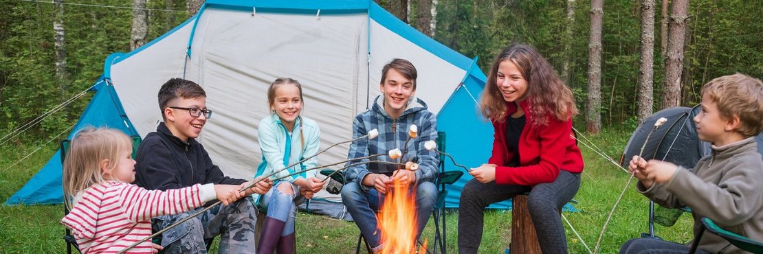 Eine Menschengruppe sitzt vor einem Lagerfeuer. Im Hintergrund steht ein Zelt im Wald.