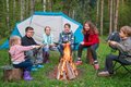 Eine Menschengruppe sitzt vor einem Lagerfeuer. Im Hintergrund steht ein Zelt im Wald.