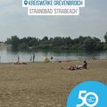 Das Bild zeigt einen Sandstrand am Strandbad „StraBeach“ in Grevenbroich.  Mehrere Personen entspannen sich am Strand, einige baden im Wasser.  Im Hintergrund ist ein See und Bäume zu sehen.  Das Bild ist hell und sonnig und vermittelt einen entspannten, sommerlichen Eindruck.  Ein Logo mit der Aufschrift „50 Jahre | 50 Orte“ und dem Hinweis auf den Rhein-Kreis Neuss ist im unteren Bereich des Bildes zu sehen. © Kreiswerke Grevenbroich GmbH
