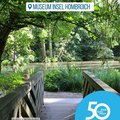 Das Bild zeigt eine Holzbrücke, die über einen kleinen Teich in einem grünen Park führt. Im Hintergrund ist üppige Vegetation und ein ruhiges Gewässer zu sehen.  Oben im Bild befindet sich ein Hinweis auf das Museum Insel Hombroich.  Rechts unten ist ein Logo mit der Aufschrift „50 Jahre | 50 Orte“ des Rhein-Kreises Neuss zu sehen. Das Gesamtbild vermittelt Ruhe und Naturverbundenheit. © Museum Insel Hombroich / Jennifer Eckert