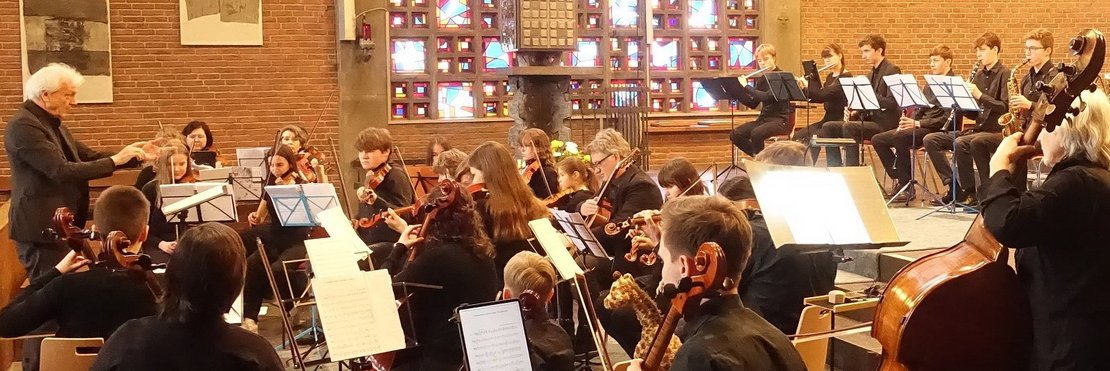 Das Bild zeigt ein Orchester, das in einer Kirche spielt. Im Vordergrund befinden sich hauptsächlich Streicher, im Hintergrund sind auch Bläser zu sehen. Ein Dirigent steht vor dem Orchester und dirigiert. Die Musiker sitzen auf Stühlen und spielen ihre Instrumente. Die Kirche hat eine Backsteinarchitektur und ein großes Buntglasfenster. Insgesamt wirkt die Szene ruhig und konzentriert.