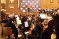 Das Bild zeigt ein Orchester, das in einer Kirche spielt. Im Vordergrund befinden sich hauptsächlich Streicher, im Hintergrund sind auch Bläser zu sehen. Ein Dirigent steht vor dem Orchester und dirigiert. Die Musiker sitzen auf Stühlen und spielen ihre Instrumente. Die Kirche hat eine Backsteinarchitektur und ein großes Buntglasfenster. Insgesamt wirkt die Szene ruhig und konzentriert.