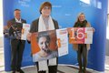 Die in der Bildbeschreibung genannten Personen stehen mit Abstand vor einem blauen Fotohintergrund und halten Kampagnen-Plakate hoch. 