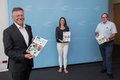 Landrat Hans-Jürgen Petrauschke, Katharina Beckmann und Robert Abts stehen mit Abstränden vor einer hellbauen Fotowand und halten Broschüren in den Händen. 